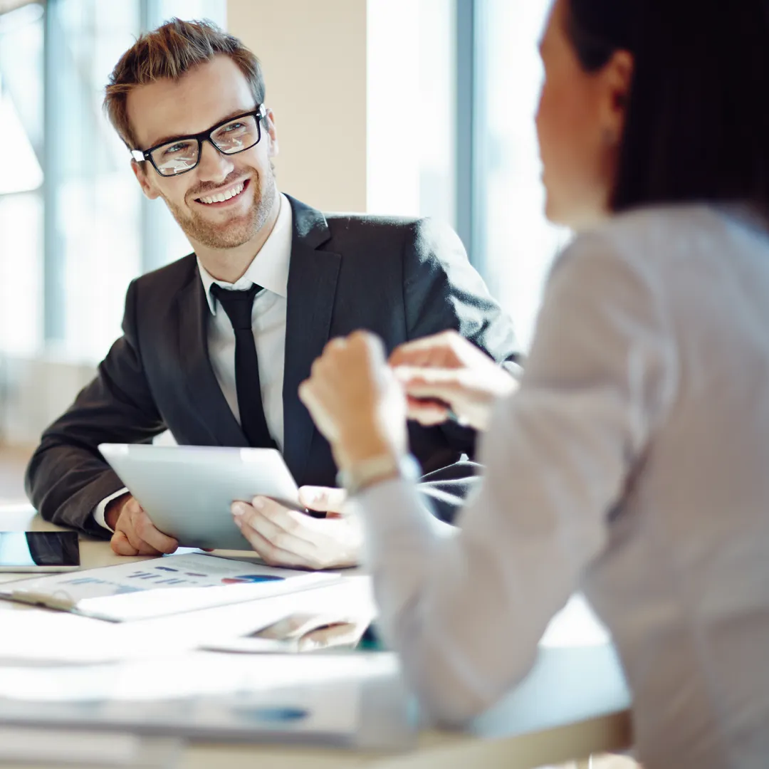 Smiling man in glasses holding a tablet during a meeting with a woman in an office.