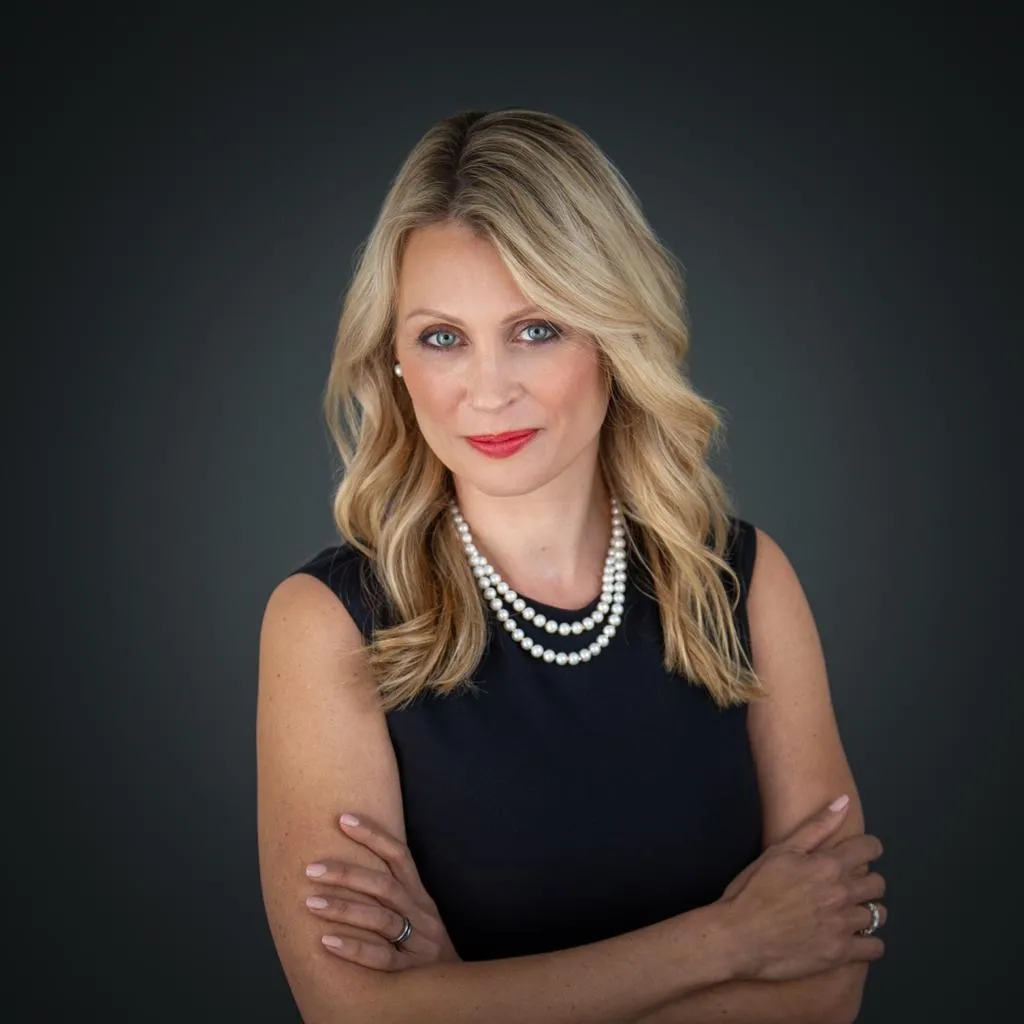 Blonde woman with red lipstick wearing pearl necklace and black sleeveless top, arms crossed.
